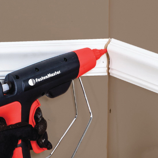A gloved hand holds a FastenMaster cordless hot air gun with a red and black body, applying white decorative molding trim to a beige wall corner.