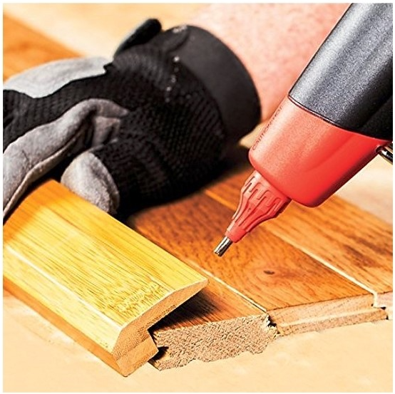 A close-up action shot of a red and black heat gun applying adhesive to a wooden tongue-and-groove panel, with a black and gray work glove holding the wood piece in place on a wooden floor.