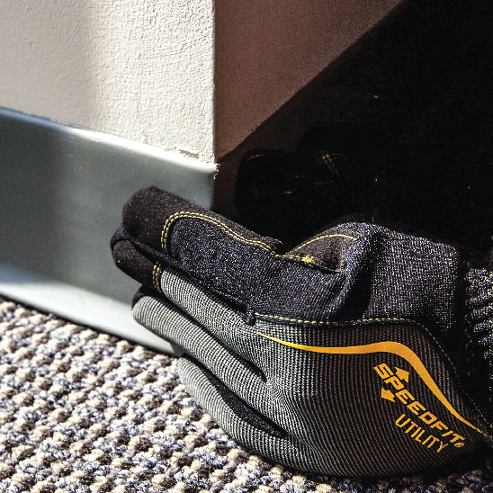 A close-up of a black and gray work glove with yellow stitching and a yellow SPEEDFIT UTILITY logo, resting on a textured carpet next to a wall baseboard.