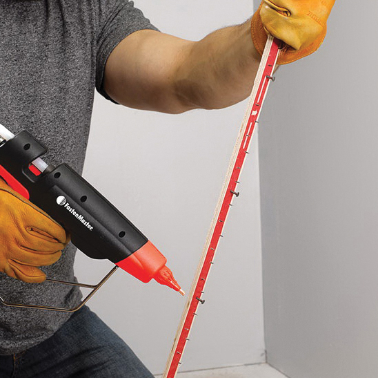 A person wearing yellow work gloves uses a black and red Fasten Master hot glue gun to apply adhesive to a long red strip against a plain light gray background.